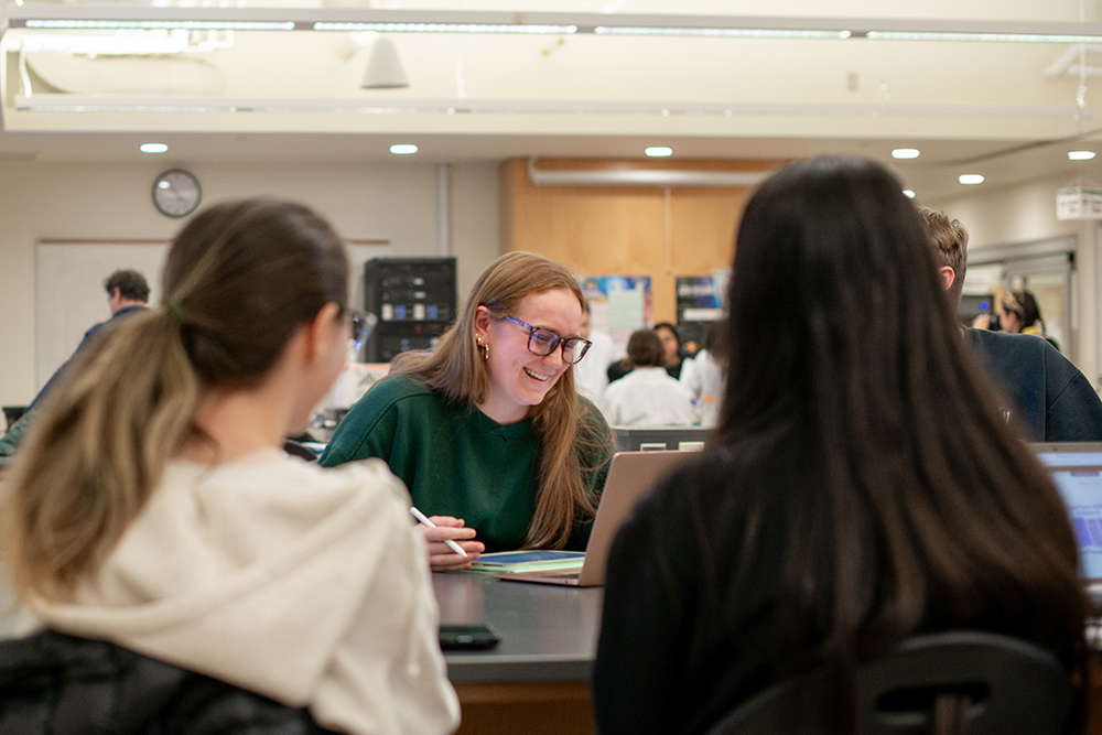 A group of students sitting together in a classroom, collaborating on laptops and notes, with one student smiling and leading the discussion.