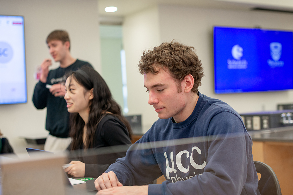 Students working in a modern classroom with laptops open; one student focuses on his screen while others engage in conversation, with Schulich Medicine & Dentistry branding visible on the monitors.