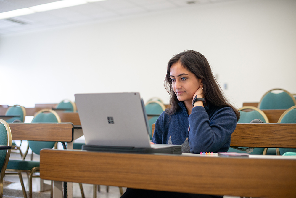 Student sitting alone in a lecture hall, focused on her laptop while studying, with rows of empty desks and green chairs around her.