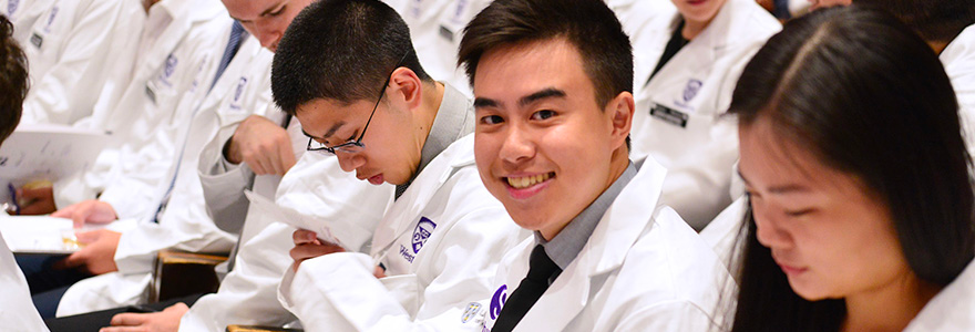 Medical students in white coats at a ceremony, one smiling at the camera.