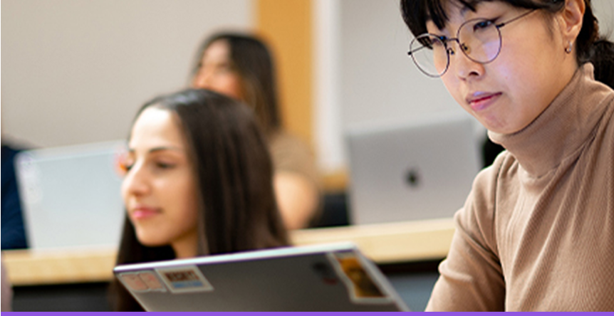 Student in glasses taking notes on a laptop in lecture hall