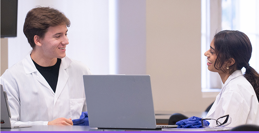 Two students in lab coats talking at a laptop.