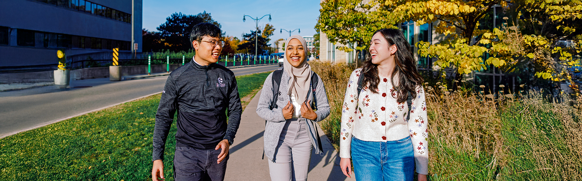 Five students sitting outdoors on a stone ledge surrounded by trees, laughing and talking together; some wearing white lab coats and carrying backpacks.