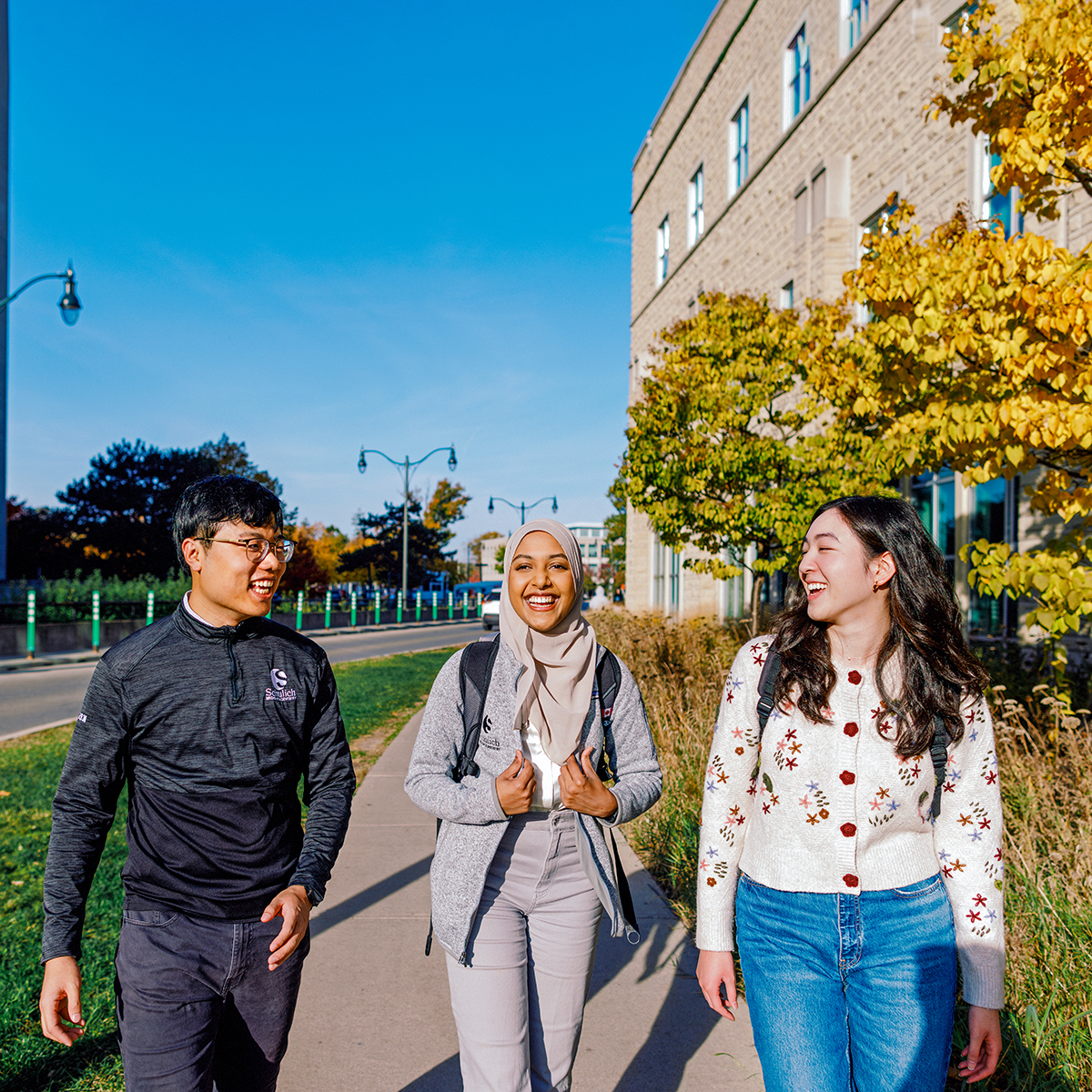 Five students sitting outdoors on a stone ledge surrounded by trees, laughing and talking together; some wearing white lab coats and carrying backpacks.