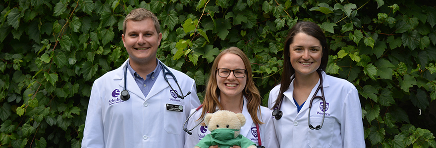 Photograph of three medicine students wearing white coats and holding a teddy bear