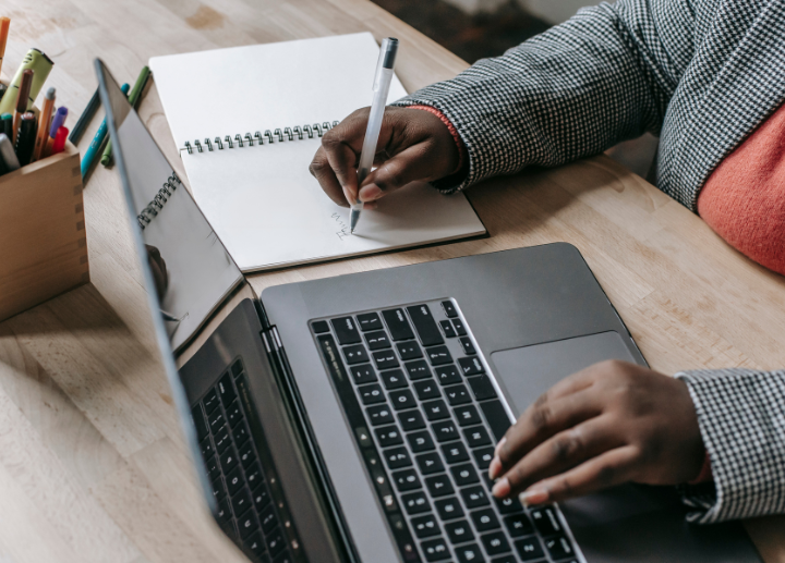 Person writing in a notebook and typing on a laptop, sitting at a desk