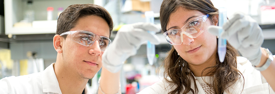 Two students wearing safety goggles and gloves closely examining test tubes in a brightly lit laboratory.