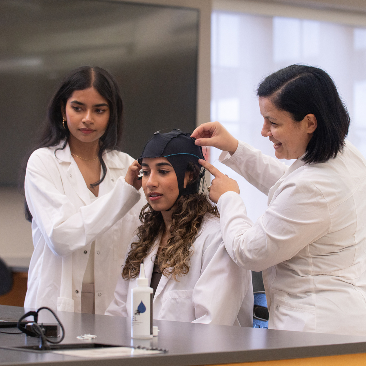 Three women in lab coats working together in a research lab; one sits while the others help her adjust an EEG cap for a neuroscience experiment, smiling and focused on the task.