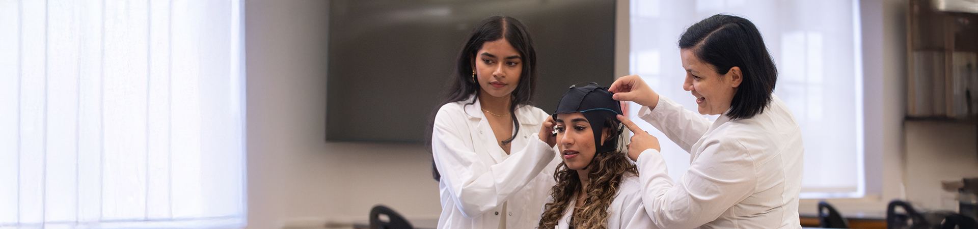 Three women in lab coats working together in a research lab; one sits while the others help her adjust an EEG cap for a neuroscience experiment, smiling and focused on the task.