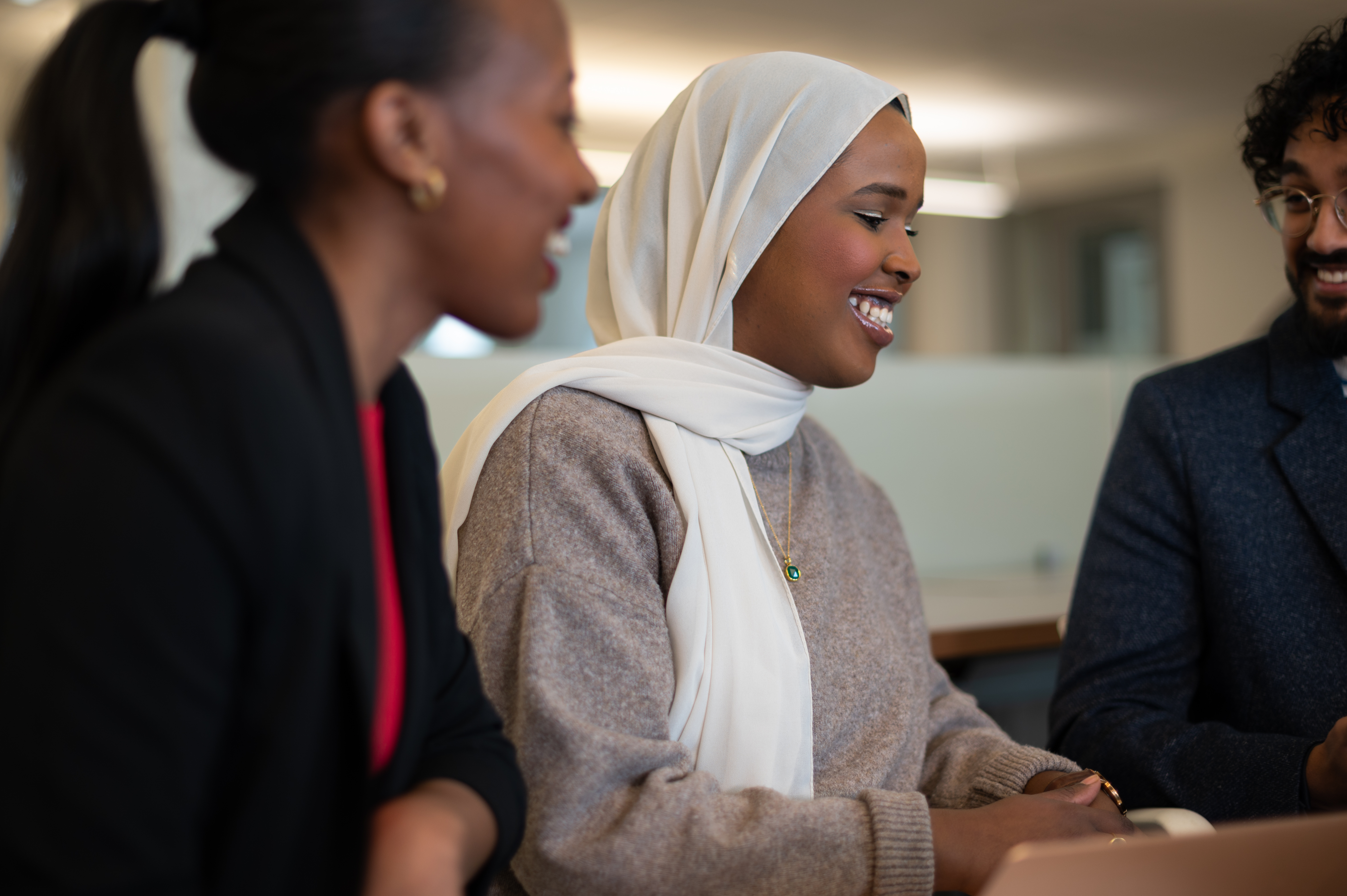 Three colleagues in conversation, smiling and collaborating at a meeting table, with one person wearing a white headscarf and light sweater.