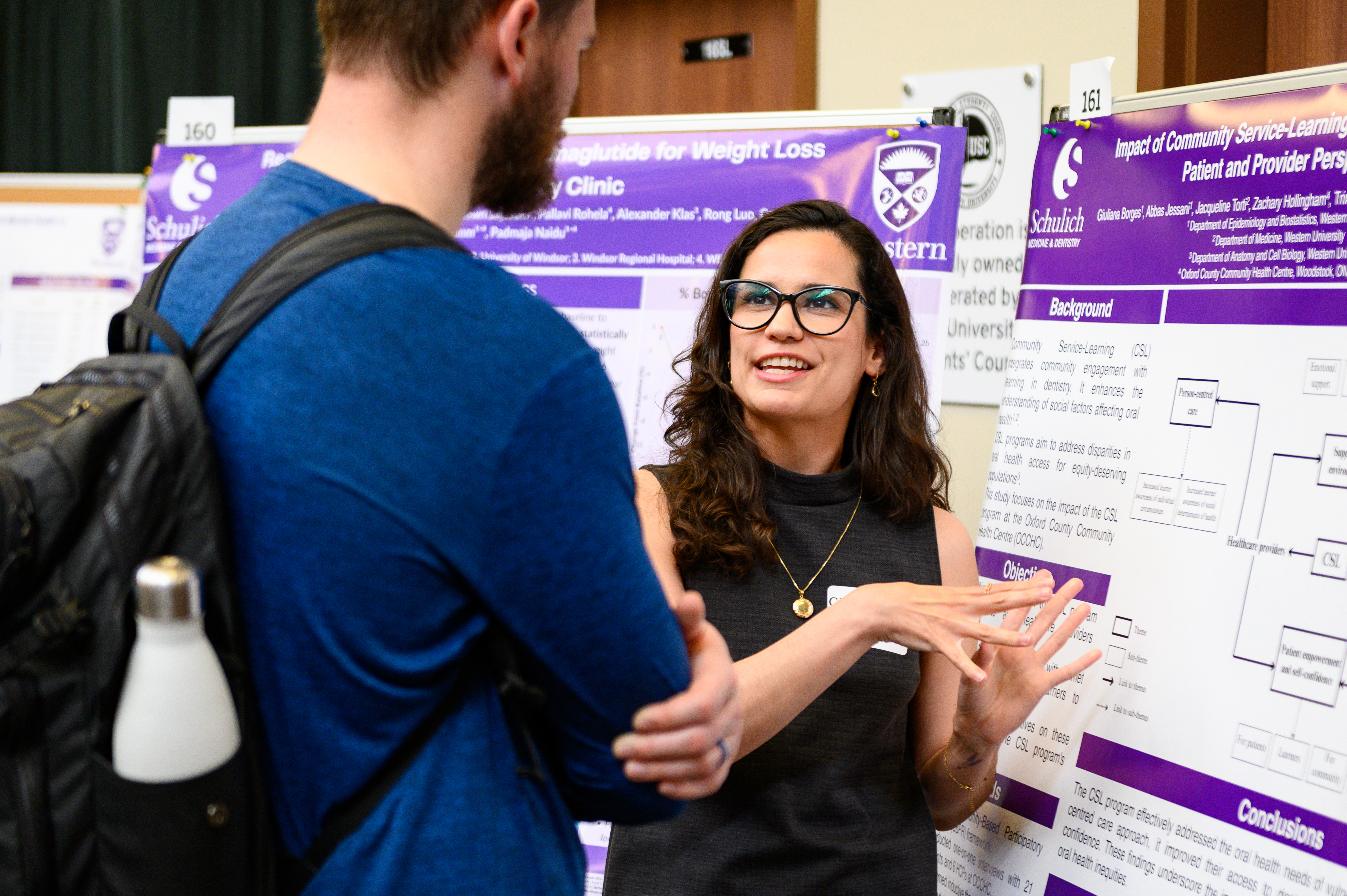 A presenter discussing her research poster with an attendee during a conference, gesturing while explaining her findings in front of a purple Schulich Medicine & Dentistry display.