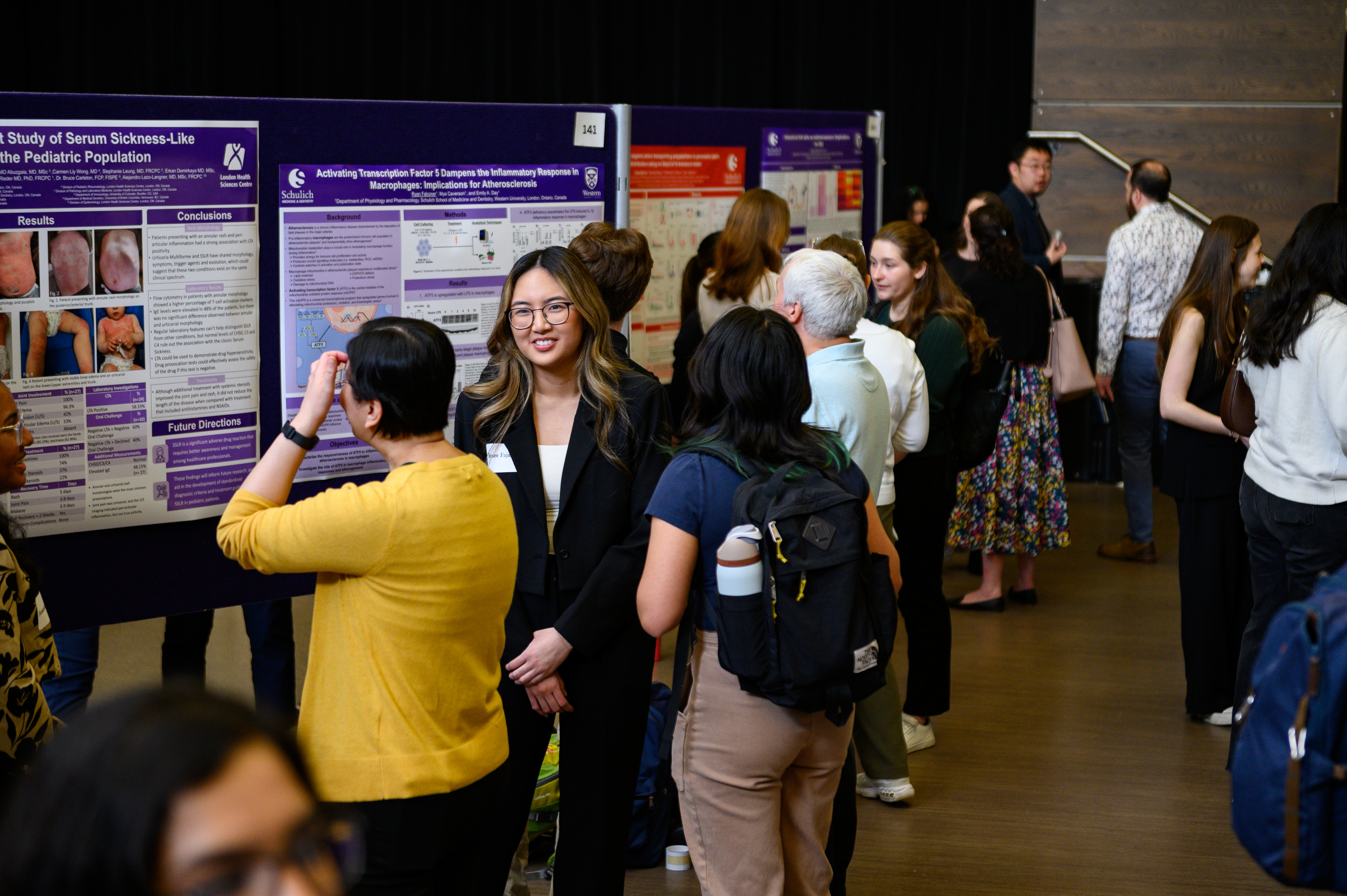A busy poster session with attendees interacting, discussing research, and viewing presentations in a large hall filled with purple posters.