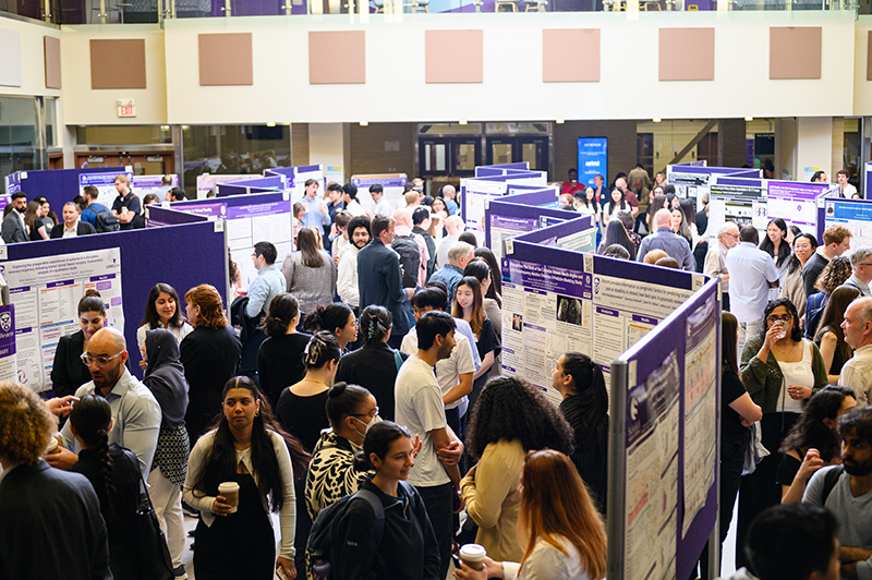 A speaker presenting at a podium during London Health Research Day, addressing an audience seated in a lecture hall with a large event banner beside the stage.