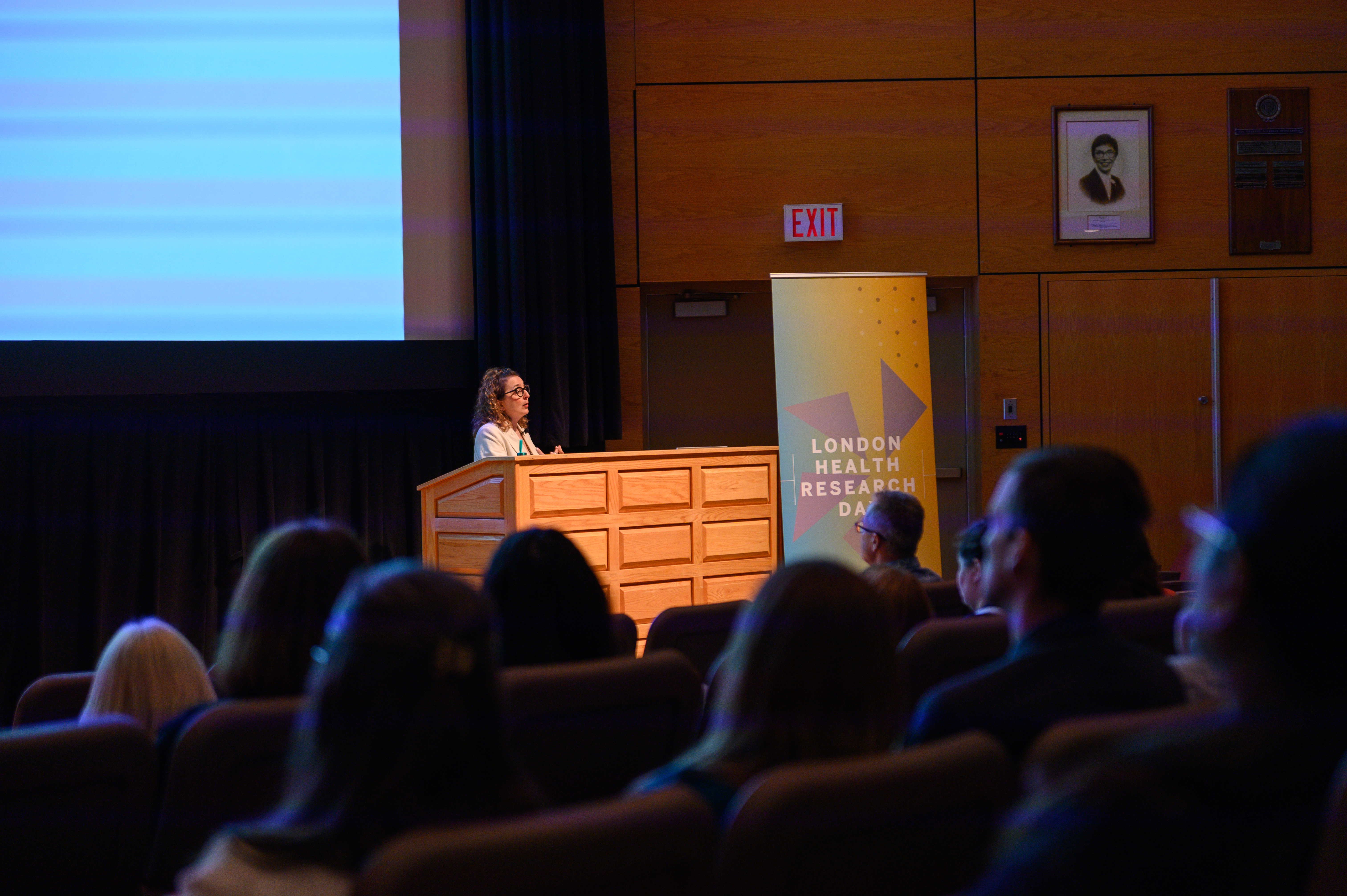 A speaker presenting at a podium during London Health Research Day, addressing an audience seated in a lecture hall with a large event banner beside the stage.