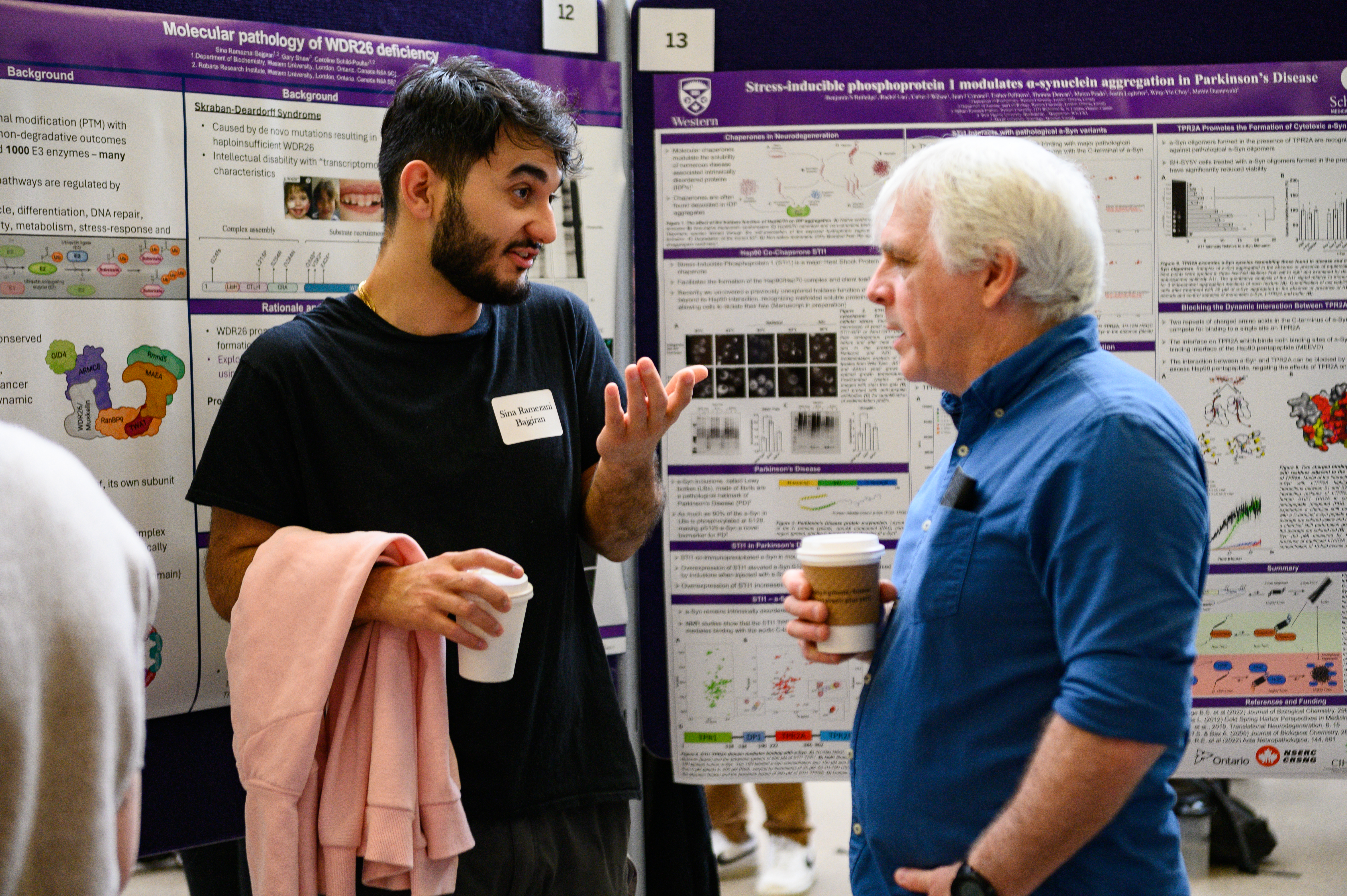 Two people holding coffee cups engaged in conversation in front of research posters at London Health Research Day, surrounded by colorful scientific displays.