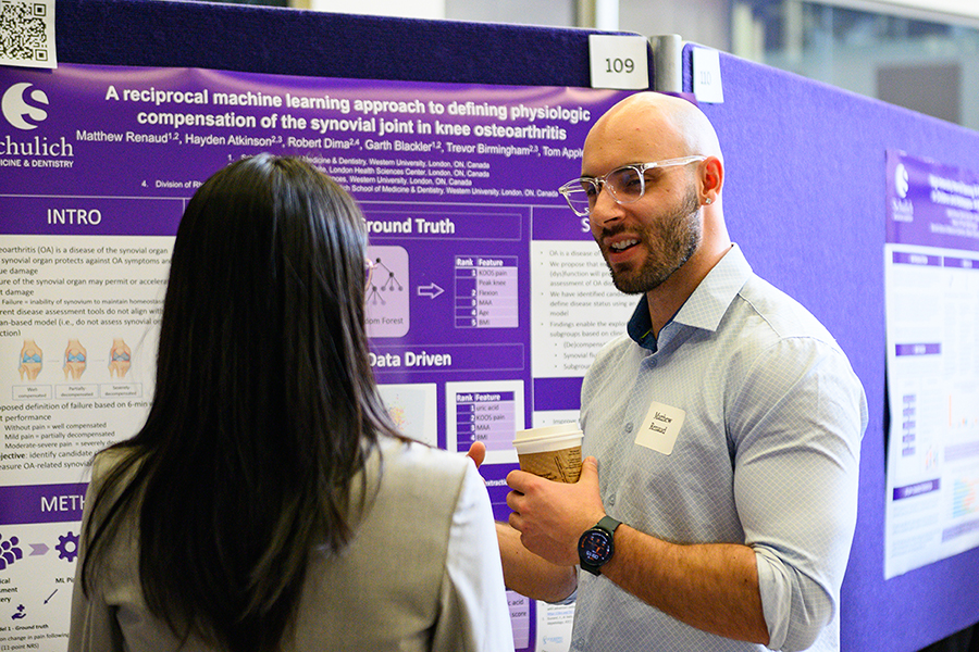 A researcher discussing a scientific poster presentation with another attendee at a conference, holding a coffee cup and standing in front of a purple Schulich Medicine & Dentistry research poster.