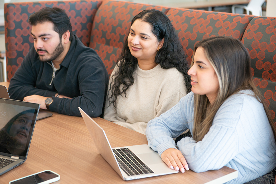 Three students sitting together in a study space with laptops open, focused and listening during a group meeting.