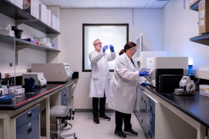 Two researchers in lab coats working in a laboratory; one operates equipment while the other adjusts a sample under bright lab lighting.