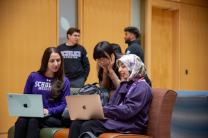 Three students sitting together indoors, smiling and working on laptops, wearing Schulich Medicine & Dentistry apparel, with two other students talking in the background.