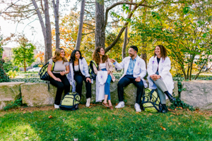 A group of five students sitting outdoors on a stone ledge surrounded by trees, chatting and smiling; some are wearing white lab coats and carrying backpacks.