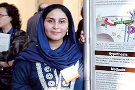 A presenter standing beside a scientific research poster, smiling at the camera, with other attendees visible in the background.