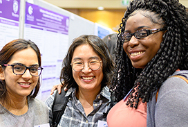 Three smiling participants standing together at a research poster session, with purple posters visible in the background.
