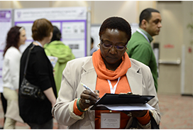 A person writing on a clipboard during a research conference, with other attendees and posters visible behind them.