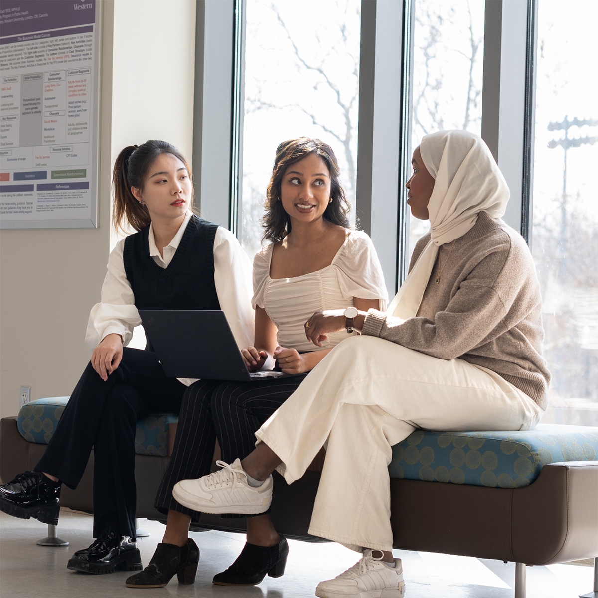 Three students sitting by large windows with sunlight streaming in, collaborating on a laptop and talking; research posters hang on the wall behind them.