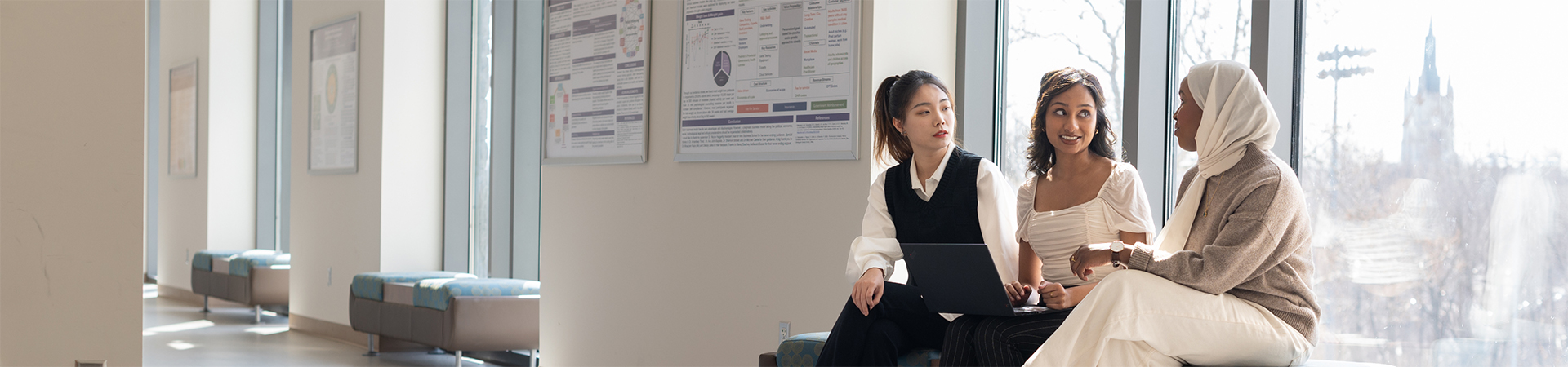 Three students sitting by large windows with sunlight streaming in, collaborating on a laptop and talking; research posters hang on the wall behind them.