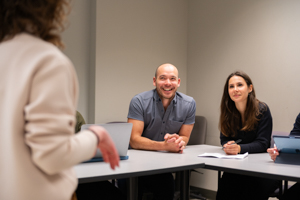 A student speaking at the front of a classroom while two instructors sit at a table smiling and listening attentively.