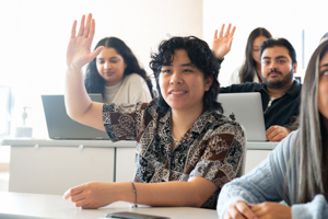 A student in a classroom raising their hand to participate, while other students with laptops listen in the background.