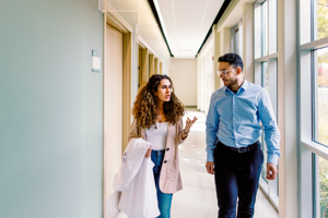 Two people walking and conversing in a bright hallway, one holding a white lab coat.