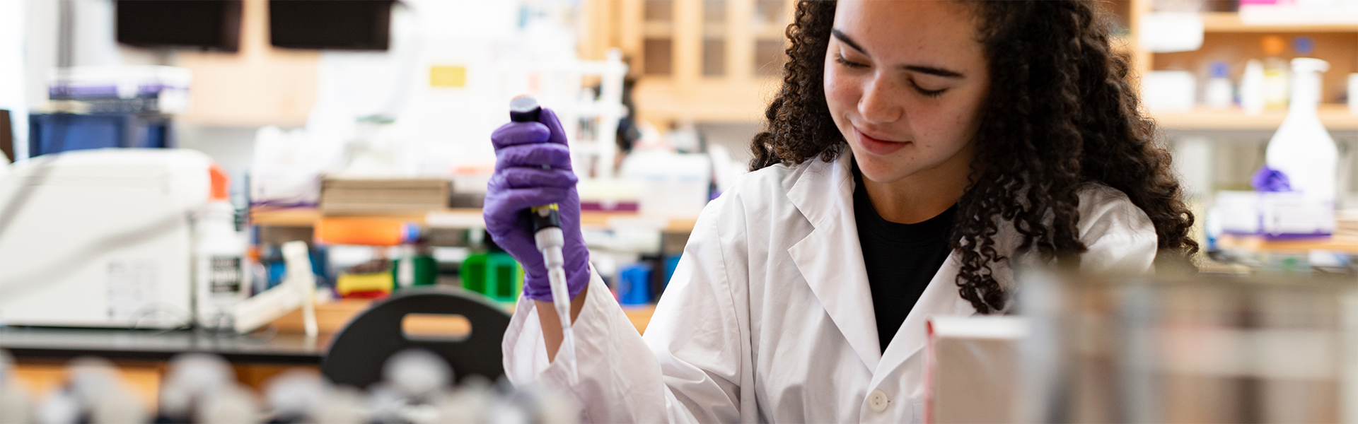 A student in a white lab coat and purple gloves pipetting samples in a laboratory, with lab equipment and supplies in the background.