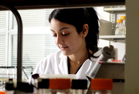 A student in a white lab coat working in a laboratory, surrounded by bottles and scientific equipment.