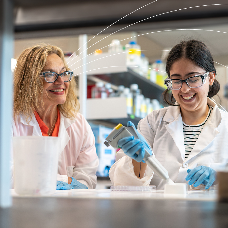 Two researchers working together in a laboratory, smiling as one conducts an experiment with a pipette