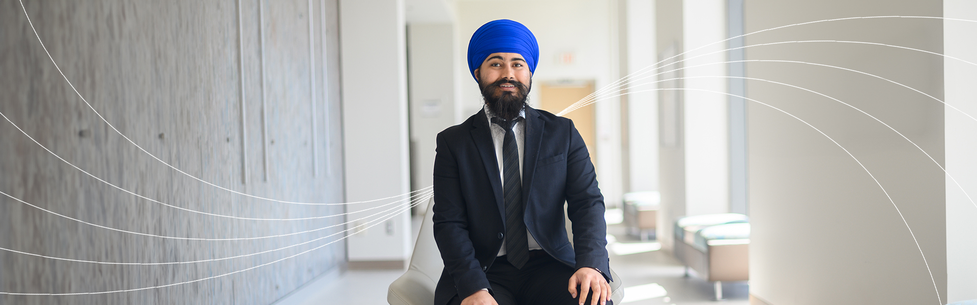 A person in a blue turban and dark suit smiling while seated in a bright hallway, with curved white graphic lines radiating across the image background.