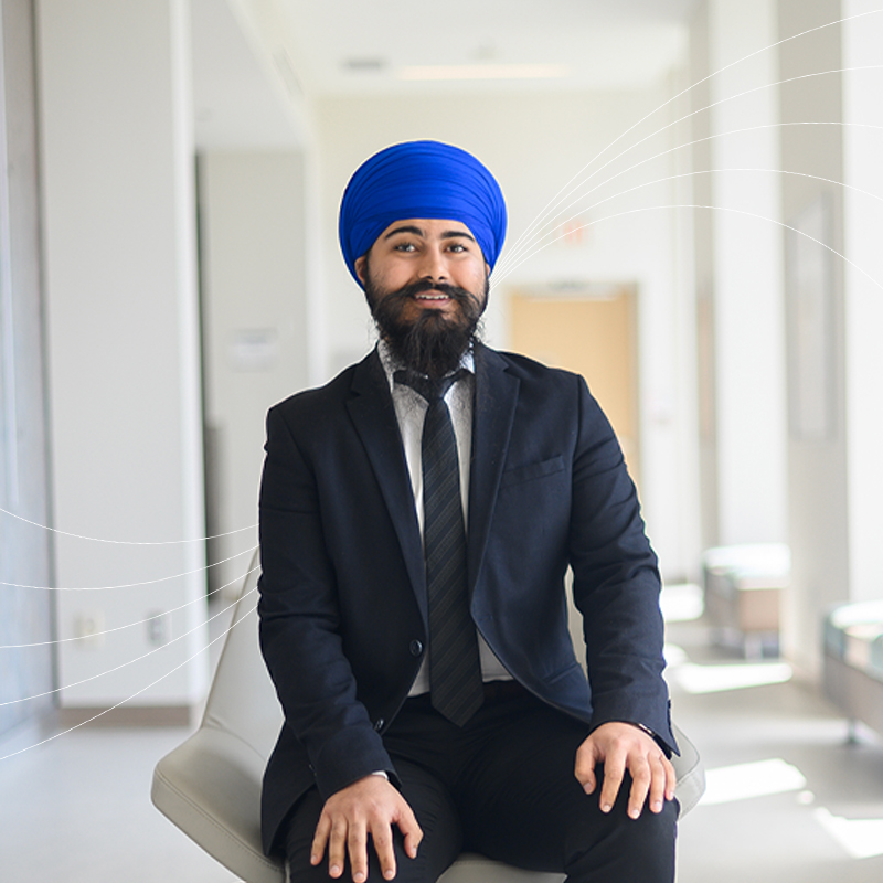 A person in a blue turban and dark suit smiling while seated in a bright hallway, with curved white graphic lines radiating across the image background.
