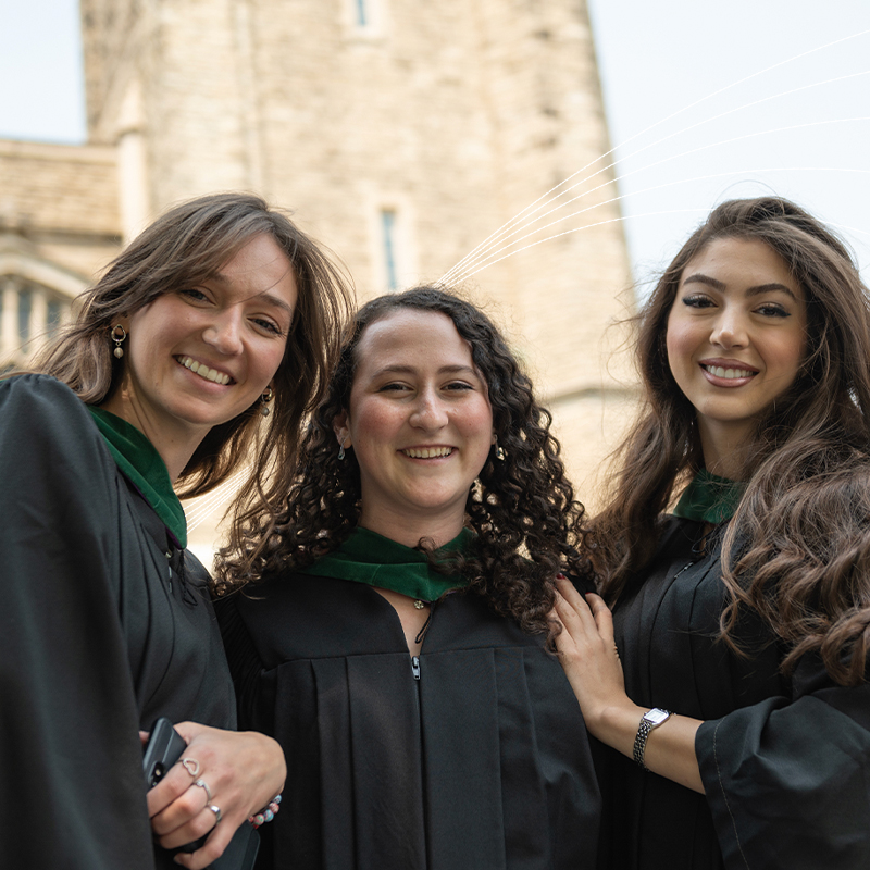 Three graduates celebrate convocation in black gowns and green hoods outside Western University.