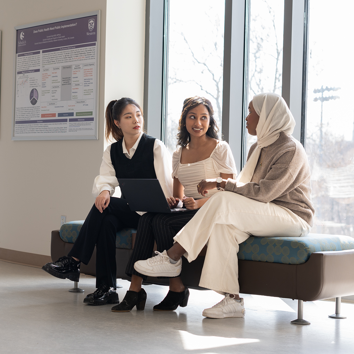 Three students sitting together by a window, talking and working on a laptop.