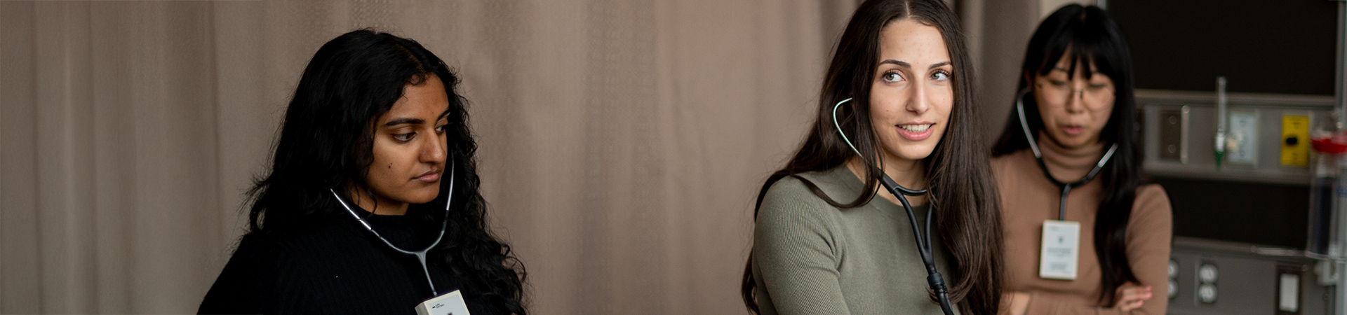 Three medical students practice using stethoscopes in a clinical setting.