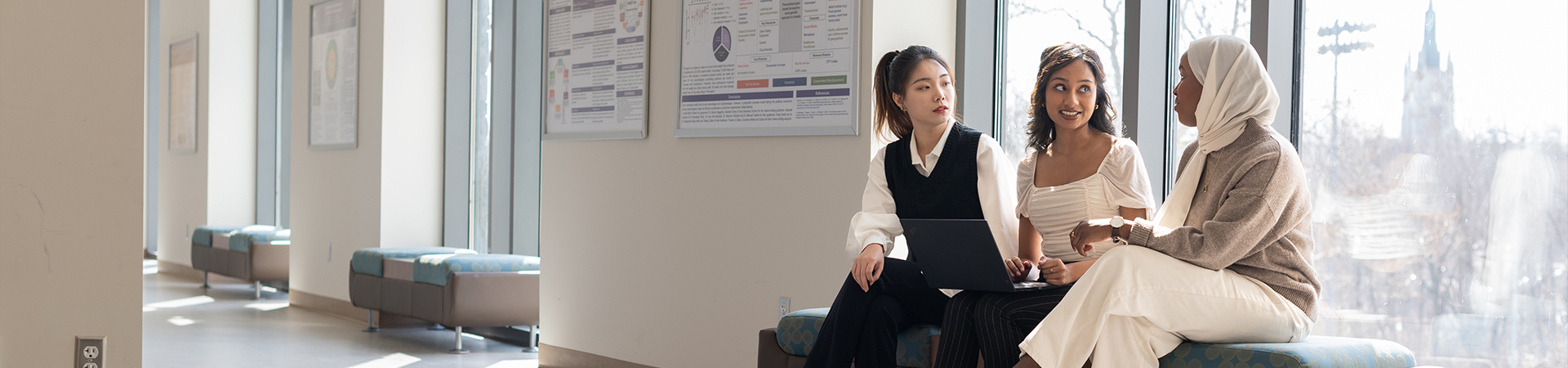 Three students sitting together by a window, talking and working on a laptop.