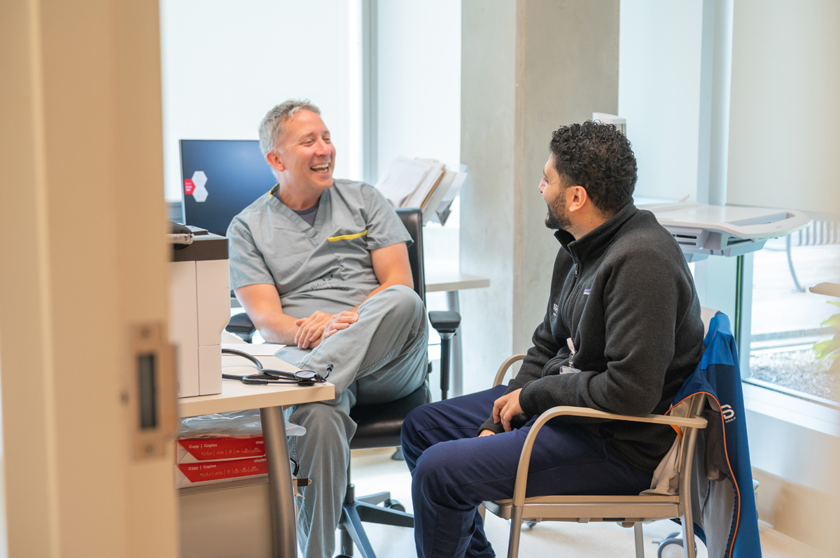 Doctor in scrubs chatting with a seated patient in an exam room.