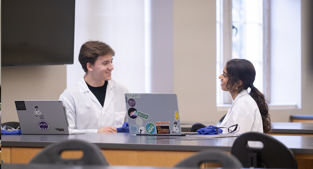 Two students discussing coursework at a classroom table with laptops