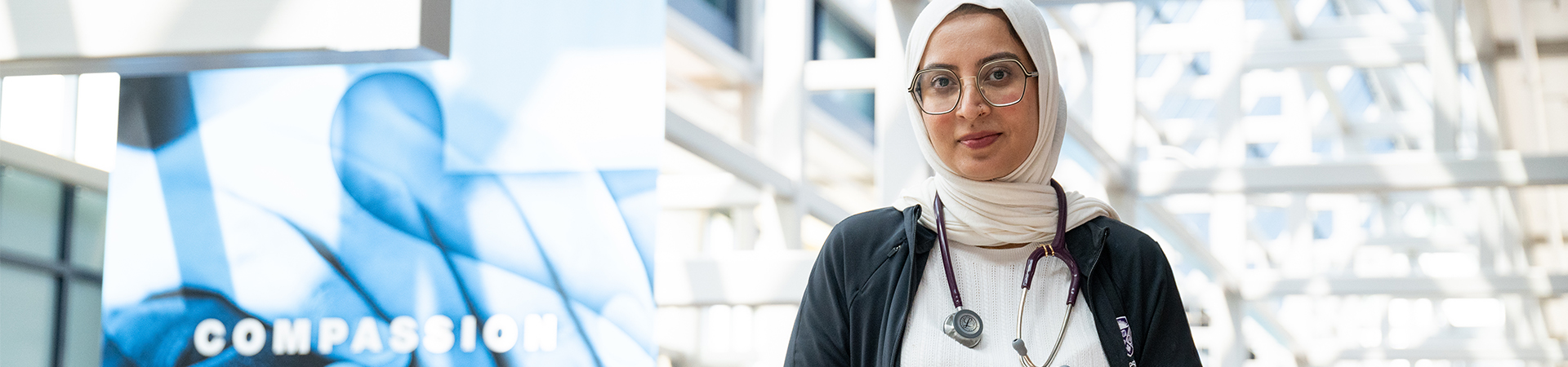 A medical trainee with a stethoscope around her neck stands in a bright, glass-roofed atrium