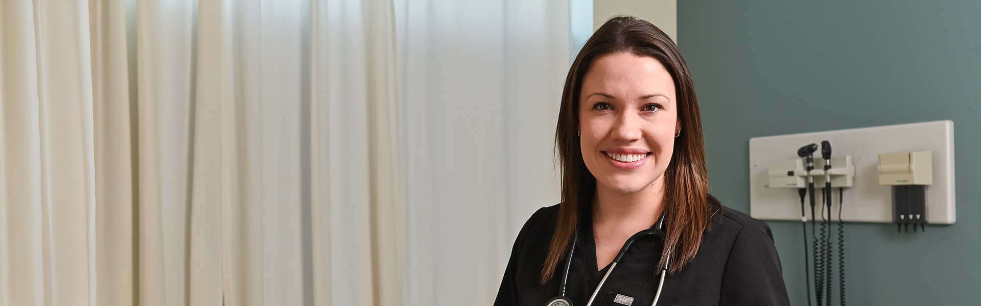 a resident standing in a hospital, smiling, wearing black scrubs and stethoscopes.