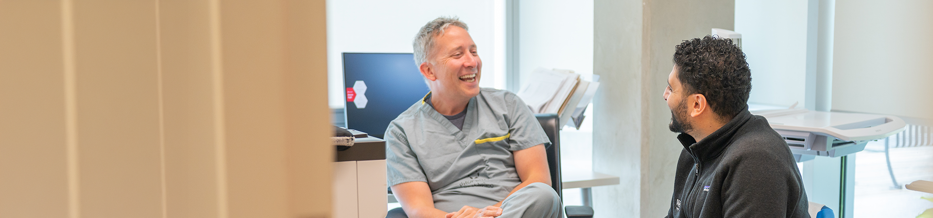 Two healthcare colleagues sitting and laughing together during a relaxed conversation in an office.