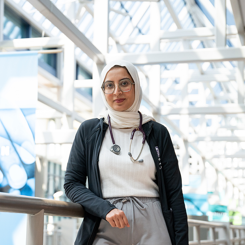 A medical trainee with a stethoscope around her neck stands in a bright, glass-roofed atrium