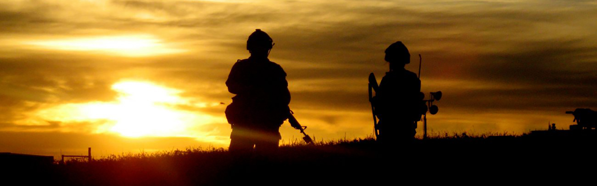 Silhouettes of two soldiers in helmets and gear standing in a field at sunset.